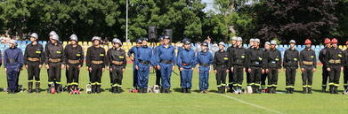 Grupa osób w różnych mundurach stojących w rzędzie na trawiastym terenie, prawdopodobnie służby mundurowe podczas uroczystości.