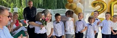 Grupa dzieci w białych koszulach i czarnych muszkach stoi w rzędzie na zewnątrz, a jedno wręcza dorosłej kobiecie bukiet kwiatów; w tle balony i liczba 20.