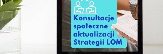 Tablet stoi na biurku z grafiką informującą o konsultacjach społecznych aktualizacji Strategii LOM, w tle widoczna roślina i część ręki osoby.