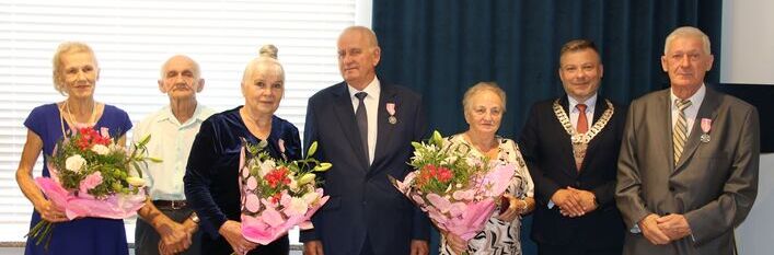 Grupa starszych osób z kwiatami oraz dwóch mężczyzn w garniturach stojących w pomieszczeniu; niektórzy odznaczeni medalami.