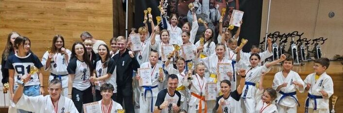 Grupa osób w kimono karate stoi na sali gimnastycznej, trzymając trofea i medale, z banerem zawodów w tle.