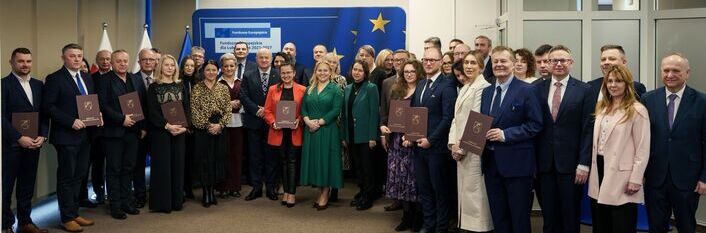 Grupa ludzi w formalnych ubraniach stoi w sali konferencyjnej. Niektórzy trzymają dokumenty. W tle widoczna jest europejska flaga i tablica informacyjna.