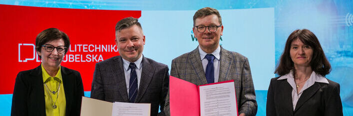 Cztery osoby stoją obok siebie, dwie trzymają dokumenty. W tle widoczne logo Politechniki Lubelskiej na czerwonym tle. Wszyscy ubrani są formalnie, w garnitury i marynarki.