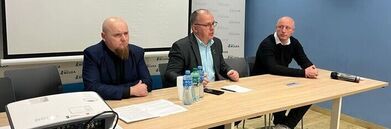 Trzech mężczyzn siedzi za stołem podczas konferencji. Na stole leżą dokumenty i butelki z wodą. W tle znajduje się ekran projekcyjny i baner. Pomieszczenie ma niebieskie ściany.