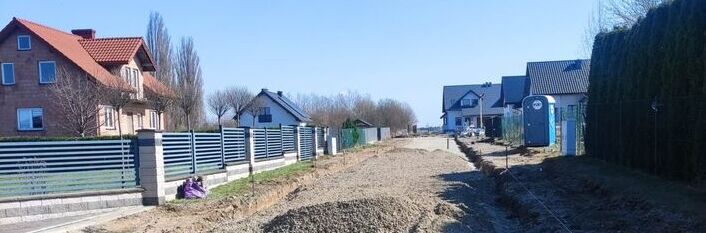 Droga w budowie wśród domów jednorodzinnych. Powierzchnia pokryta żwirem, po bokach ogrodzenia i trawniki. Na końcu widoczne niebo i drzewa, po prawej toitoi.