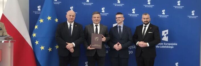 Na zdjęciu czterech mężczyzn w garniturach stoi na tle niebieskiej ścianki z logo funduszy europejskich i flaga Polski oraz Unii Europejskiej. Jeden z mężczyzn trzyma dokument. Wszyscy się uśmiechają.
