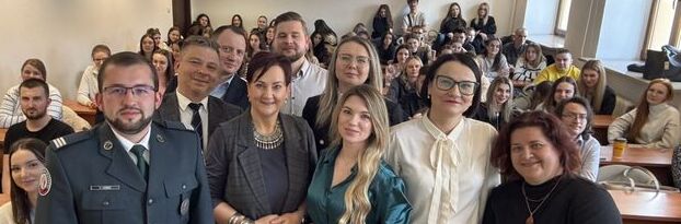 Na zdjęciu znajduje się grupa ludzi w sali wykładowej. Z przodu stoi osiem osób, w tym jedna w mundurze. W tle siedzi większa liczba słuchaczy przy stołach. Atmosfera jest formalna i edukacyjna.