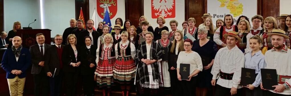 Grupa osób w tradycyjnych strojach ludowych pozuje na podium w pomieszczeniu urzędowym. W tle widoczne są herby i mapa Polski. Niektóre osoby trzymają dokumenty.