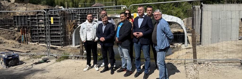 Grupa mężczyzn stoi przed placem budowy mostu, otoczona metalowym ogrodzeniem. W tle widoczne są maszyny budowlane oraz drzewa. Pogoda jest słoneczna, na niebie kilka chmur.