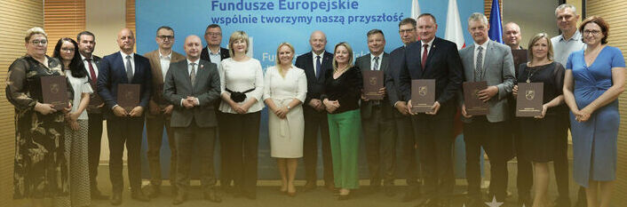 Na zdjęciu grupa osób stoi w rzędzie, trzymając tablice. Za nimi widoczny jest baner z napisem "Fundusze Europejskie wspólnie tworzymy naszą przyszłość". W tle znajdują się flagi i symbol Unii Europejskiej.