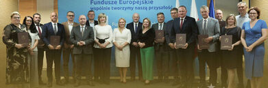 Na zdjęciu grupa osób stoi w rzędzie, trzymając tablice. Za nimi widoczny jest baner z napisem "Fundusze Europejskie wspólnie tworzymy naszą przyszłość". W tle znajdują się flagi i symbol Unii Europejskiej.