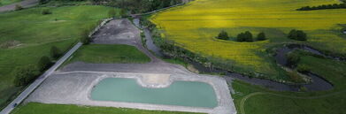 Panorama wiejskiego krajobrazu z dronowej perspektywy. W centrum: mały zbiornik wodny otoczony polami, w tym rzepakowymi. Widoczne są także drogi, mostek i rozległa wiejska zabudowa w tle.