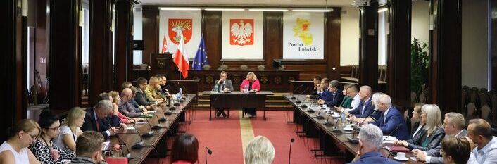 Sala konferencyjna z dużym stołem w kształcie podkowy, wokół którego siedzi wiele osób. Z przodu widoczny stół prezydialny z trzema osobami. W tle flagi i logo powiatu.