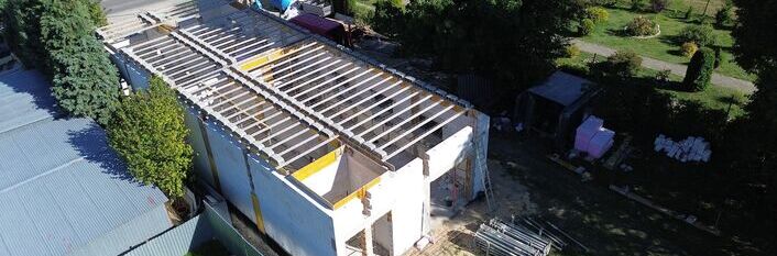 Budynek w trakcie budowy widziany z lotu ptaka. Konstrukcja ma zainstalowany dach szkieletowy. Wokół widoczne są materiały budowlane i drzewa. Budowa znajduje się na zielonym terenie w pobliżu drogi.