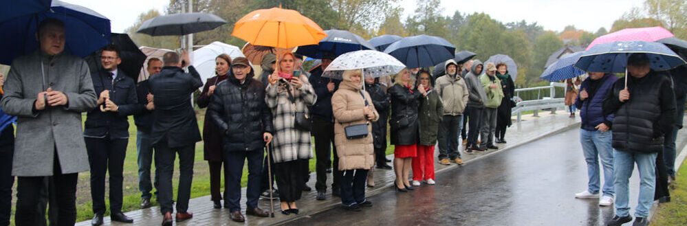 Grupa ludzi stoi na mokrej ulicy, trzymając kolorowe parasole. Ubrani w płaszcze, chronią się przed deszczem. W tle drzewa w jesiennym krajobrazie.