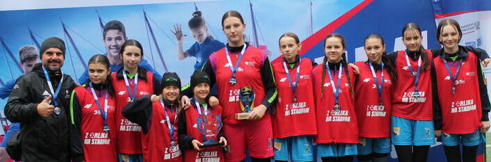 Dziesięć osób w kolorowych strojach sportowych pozuje na tle baneru ze zdjęciami młodych piłkarzy. Większość z nich nosi czerwone kamizelki, a jedna osoba trzyma piłkę. Wszyscy wydają się być zadowoleni.