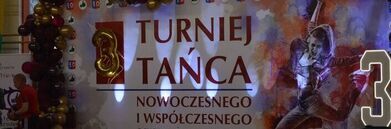 Na zdjęciu widać dekoracje turnieju tańca nowoczesnego i współczesnego, z dużym banerem i balonami. Na banerze znajduje się sylwetka tancerza. Wystrój jest kolorowy i przyciąga uwagę.