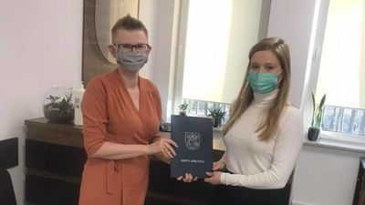 Dwie kobiety w maseczkach trzymające granatową okładkę z herbem Gminy Jabłonna, stoją na tle okien.