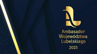 Złote logo z napisem "Ambasador Województwa Lubelskiego 2023" na ciemnoniebieskim tle z abstrakcyjnym, złotym elementem po lewej stronie.