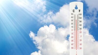 Na zdjęciu termometr pokazuje wysoką temperaturę, wskazując na skali stopnie Celsjusza i Fahrenheita. W tle widoczne są białe chmury i niebieskie niebo, przez które przeświecają promienie słoneczne.