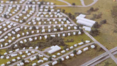Zdjęcie przedstawia makietę osiedla z małymi, białymi budynkami. Dookoła nich widać siatkę dróg i zielone tereny. Układ przypomina typowy amerykański suburb z głównymi i bocznymi ulicami.