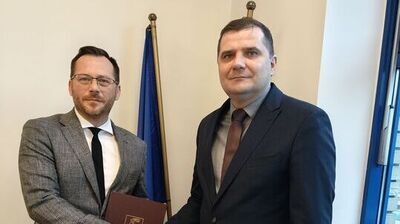 Dwóch mężczyzn w garniturach stoi w pomieszczeniu biurowym, podają sobie dłonie na powitanie lub gratulacje. Jeden z nich trzyma brązową teczkę z napisem „MARSZAŁEK WOJEWÓDZTWA LUBELSKIEGO”. W tle znajduje się niebieska flaga na złotym stojaku.Obaj panowie wyglądają poważnie i oficjalnie.