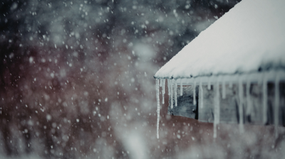 Opadające płatki śniegu, dach pokryty grubą warstwą białego śniegu i zwisające długie sople lodu. Scena przedstawia zimowy krajobraz, rozmyte tło dodaje atmosfery chłodu i ciszy.
