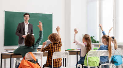 Sala lekcyjna z nauczycielem stojącym przy tablicy i grupą uczniów siedzących przy ławkach. Kilkoro dzieci podnosi ręce, chcąc odpowiedzieć na pytanie nauczyciela. Na krzesłach mają zawieszone kolorowe plecaki. W tle widoczna jest zielona tablica i jasne ściany, a światło wpada przez duże okno. Pomieszczenie wygląda przestronnie i uporządkowanie.
