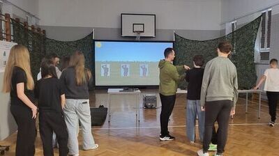 Grupa osób stoi w sali gimnastycznej przed ekranem, na którym wyświetlany jest symulator strzelnicy z tarczami. Na ścianach zawieszone są siatki maskujące. Osoby są w trakcie ćwiczeń lub instruktażu.