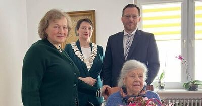 Zdjęcie przedstawia cztery osoby w jasnym pokoju. Na pierwszym planie, na krześle po prawej, siedzi starsza kobieta z siwymi włosami spiętymi z tyłu, ubrana w niebieski sweter i ciemną spódnicę. Trzyma w ramionach duży bukiet różowych i czerwonych kwiatów owinięty różowym papierem.

Za nią stoją trzy osoby:
Kobieta po lewej w zielonym swetrze, krótkie jasne włosy, patrzy w stronę aparatu.
Kobieta w środku w zielonym żakiecie, nosi łańcuch oficjalny wokół szyi (wygląda jak symbol urzędu), ma krótko ciemne włosy.
Mężczyzna po prawej w ciemnym garniturze, z krawatem w kratę i okularami.

W tle: jasne ściany, obraz w ramie za środkową osobą, po prawej okno z żółto-białą roletą i kilka doniczkowych roślin na parapecie. Na podłodze drewniana powierzchnia i fragment ozdobnego dywanu; po prawej widać stół z obrusem. Osoby wyglądają na zebrane przy uroczystej okazji; atmosfera spokojna i formalna.