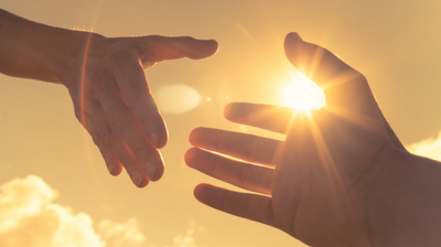 Dwie ludzkie dłonie wyciągnięte ku sobie na tle zachodzącego lub wschodzącego słońca, którego promienie przeświecają między palcami.