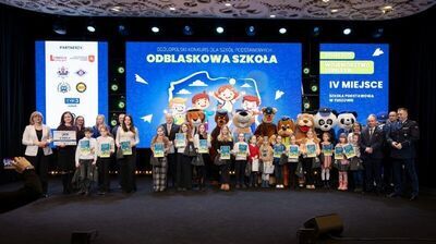 Grupa kilkudziesięciu osób stojących na scenie z dyplomami w rękach.
W środku rzędu stoją dzieci i nastolatki, większość trzyma kolorowe dyplomy i ma odświętne stroje – koszule, swetry, spódnice i spodnie materiałowe.
Pomiędzy dziećmi w kilku miejscach stoją duże maskotki w kostiumach zwierząt i postaci bajkowych, między innymi pies, panda, miś i inne pluszowe postacie.
Po lewej stronie stoi kilka dorosłych kobiet, jedna trzyma dużą tabliczkę z napisem nagrody pieniężnej.
Po prawej stronie stoi kilku dorosłych mężczyzn w garniturach i w mundurach policyjnych.
Za grupą w tle jest bardzo duży ekran LED z niebieską grafiką – na środku widać rysunkowe postacie dzieci w odblaskowych kamizelkach oraz duży napis „ODBLASKOWA SZKOŁA”, a wyżej mniejszy: „OGÓLNOPOLSKI KONKURS DLA SZKÓŁ PODSTAWOWYCH”.
Po prawej stronie ekranu jest pionowy panel z napisem „WOJEWÓDZTWO LUBELSKIE” oraz „IV MIEJSCE SZKOŁA PODSTAWOWA W TUSZOWIE”.
Po lewej stronie sceny stoi pionowa tablica z logotypami partnerów i instytucji.
Przed sceną, w lewym dolnym rogu zdjęcia, widać osobę robiącą zdjęcie telefonem skierowanym na grupę.