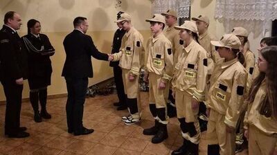 Grupa dzieci i nastolatków w jasnożółtych mundurach strażackich stoi w szeregu w ozdobionej sali.
Mundury mają czarne naszywki na ramionach i żółte pasy odblaskowe na rękawach oraz nogawkach; na piersiach widoczny jest czarny napis „STRAŻ” na żółtym tle.
Część młodzieży ma beżowe czapki z daszkiem, reszta jest bez nakrycia głowy.
Większość osób w mundurach trzyma w dłoniach czerwone materiałowe worki–plecaki.
Po lewej stronie dwóch dorosłych mężczyzn w ciemnych garniturach lub galowych mundurach rozmawia z pierwszym chłopcem w szeregu, jakby wręczali mu coś lub gratulowali.
Po prawej stronie, pod ścianą, stoi kilka dziewcząt w takich samych mundurach, a przed nimi kilkoro dzieci w codziennych ubraniach, także z czerwonymi woreczkami.
Na suficie i pod nim rozwieszona jest duża liczba balonów w kolorach złotym, czarnym, białym i czerwonym, z długimi zwisającymi wstążkami, co nadaje sali uroczysty, imprezowy charakter.