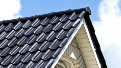 Dach dwuspadowy budynku pokryty czarną, błyszczącą dachówką ceramiczną o falistym profilu. Dachówka ułożona jest w równych rzędach, tworząc rytmiczny wzór fal. Krawędzie dachu wykończone są pasami blachy w kolorze zbliżonym do dachówki. Pod połacią widoczny jest fragment elewacji szczytowej w jasnym kolorze, z pionowym deskowaniem lub panelami. W części szczytowej znajduje się fragment okna z białą ramą o wielokątnym kształcie. W tle widoczne jest niebo z białymi obłokami.