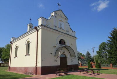 Niewielki kościół o jasnych, kremowych ścianach z ciemnoczerwonym cokołem.
Bryła jest prostokątna, jednokondygnacyjna, z łagodnie dwuspadowym dachem i trzema niewielkimi wieżyczkami zwieńczonymi metalowymi krzyżami.
Fasada frontowa ma dekoracyjny szczyt z kręconymi, symetrycznymi gzymsami oraz wnękę z białą figurą świętego umieszczoną wysoko pośrodku.
Wejście główne to drewniane, dwuskrzydłowe drzwi w wysokim, półokrągłym portalu; nad nimi znajduje się obraz religijny w zaokrąglonej wnęce.
Przed drzwiami jest przeszklone zadaszenie na metalowych, ozdobnie wygiętych wspornikach.
Boczne ściany kościoła mają wąskie, wysokie okna zakończone łukiem, ujęte w gładkie obramienia.
Wokół budynku rozciąga się zadbany, zielony trawnik i chodniki z czerwonej kostki; przed wejściem stoją drewniane ławki i niskie stoliki.
W tle widoczne są pojedyncze drzewa iglaste, ogrodzenie z czerwonej cegły i fragmenty domów jednorodzinnych pod błękitnym, bezchmurnym niebem.