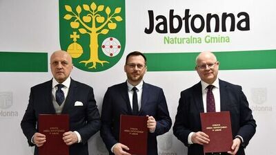 Trzech mężczyzn w garniturach trzymających bordowe teczki.
Po lewej stoi łysy mężczyzna o jasnej karnacji, w ciemnym granatowym garniturze, jasnoszarej kamizelce, białej koszuli i granatowym krawacie z wzorem; z kieszonki wystaje biały poszetka.
Pośrodku stoi mężczyzna o jasnej karnacji, z krótkimi brązowymi włosami i brodą, w granatowym garniturze, białej koszuli i ciemnym krawacie; ma okulary w cienkich oprawkach.
Po prawej jest mężczyzna o jasnej karnacji, łysy, w granatowym garniturze, białej koszuli i bordowym krawacie z wzorem; nosi okulary.
Każdy z nich trzyma przed sobą bordową teczkę z złotym napisem „DYREKTOR URZĘDU STATYSTYCZNEGO W LUBLINIE”.
W tle widoczne jest duże zielono-żółte godło z drzewem jabłoni z owocami oraz napis „Jabłonna Naturalna Gmina” na jasnym tle.