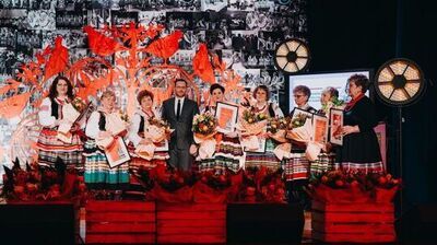 Grupa osób stojąca na scenie w trakcie uroczystości.
Na środku stoi mężczyzna w ciemnym garniturze z białą koszulą i krawatem, po obu jego stronach jest kilka kobiet w kolorowych, ludowych strojach – mają białe koszule, ciemne gorsety i długie spódnice w poziome, wielobarwne pasy.
Każda z kobiet trzyma duży bukiet kwiatów owinięty jasnym papierem oraz prostokątną ramę z dokumentem lub dyplomem w środku.
Po prawej stronie stoi jeszcze jedna kobieta w czarnej sukience z kolorowym, pasowym wykończeniem u dołu oraz czerwonym naszyjnikiem; trzyma w rękach ramę i kopertę.
Przed grupą, na przodzie sceny, ustawione są drewniane skrzynki wypełnione czerwonymi kwiatami.
W tle widać duży, dekoracyjny motyw wycinanki w intensywnym czerwonym kolorze z ptakami i roślinnymi kształtami oraz czarno‑białą ścianę z wieloma małymi zdjęciami.
Po bokach sceny stoją duże, okrągłe lampy świecące ciepłym światłem.