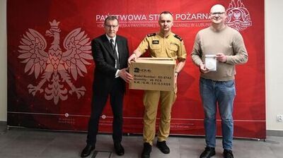 Trzech mężczyzn stojących przed czerwonym tłem z napisem „PAŃSTWOWA STRAŻ POŻARNA” oraz godłami (biały orzeł po lewej i emblemat straży po prawej). Po lewej stoi mężczyzna w czarnym garniturze, białej koszuli i czarnym krawacie, w okularach. W środku stoi mężczyzna w piaskowym mundurze strażackim, trzyma duże kartonowe pudło z nadrukiem „ELHOME”, „Model Number: SD-87A2”, „Quantity: 40 PCS”, „N.W./G.W.: 5.6/6.5 KGS”. Po prawej stoi mężczyzna w jasnym swetrze i niebieskich dżinsach, w okularach, trzyma zapakowane urządzenie z napisem „CZUJNIK DYMU” na opakowaniu