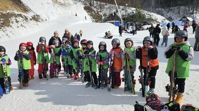 Grupa dzieci na stoku narciarskim ubrana w kaski i kolorowe kamizelki, trzymająca narty. W tle inne osoby cieszące się zimowym dniem. Śnieżny krajobraz otoczony drzewami.