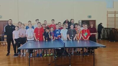 Grupa dzieci i dorosłych stojąca w sali gimnastycznej za stołem do tenisa stołowego.
Widać około dwudziestu kilku osób, głównie dzieci i nastolatków, ustawionych w dwóch rzędach.
Wiele osób ma na szyi medale na wstążkach, a kilka trzyma małe puchary.
Na pierwszym planie stoi niebieski stół do ping-ponga z siatką pośrodku, na metalowych nogach z kółkami.
Po lewej stronie stoi dorosły mężczyzna w czarnym dresie, a po prawej dorosła kobieta w ciemnym swetrze; w tle jest jeszcze kilku dorosłych.
W tle są jasne ściany sali, drabinki gimnastyczne po prawej oraz sprzęt sportowy złożony przy ścianie.
