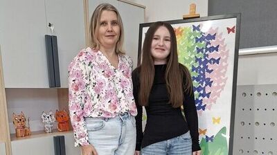 Dwie osoby stojące obok siebie w pomieszczeniu, przy sztaludze z kolorowym obrazem motyli.

Po lewej stronie stoi dorosła kobieta o jasnej cerze, blond włosach do ramion i neutralnym wyrazie twarzy. Ma na sobie jasną koszulę w różowy kwiatowy wzór, jasnoniebieskie dżinsy i białe buty sportowe.

Po prawej stoi nastolatka o jasnej cerze i długich, prostych, brązowych włosach. Ma na sobie czarną, dopasowaną bluzkę z długim rękawem, niebieskie dżinsy i jasne buty.

Za nimi, po prawej, stoi sztaluga z oprawioną pracą przedstawiającą wiele kolorowych motyli (m.in. różowe, fioletowe, niebieskie, zielone, żółte) ułożonych w gęsty wzór.

W tle po lewej widać jasne szafki; na górze stoją drewniane figurki jeleni, kolorowa zabawka z drucikami i koralikami oraz globus. Na półce są małe figurki kotów. Po prawej stronie jest fragment ściany z jasną tablicą z okrągłymi otworami i grzejnikiem.