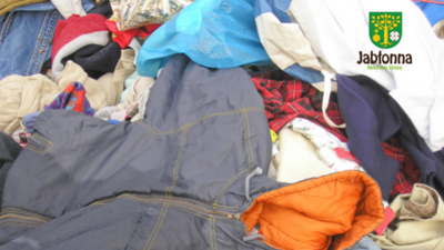 Stos ubrań i tkanin w różnych kolorach, z widocznym dużym fragmentem szarych spodni lub krótkich spodenek z żółtawymi szwami na pierwszym planie. Wokół leżą inne elementy odzieży, m.in. pomarańczowa część materiału, czerwono-czarna krata, jasne i ciemne ubrania oraz niebieska tkanina. W prawym górnym rogu znajduje się logo z herbem i napisem „Jabłonna”.