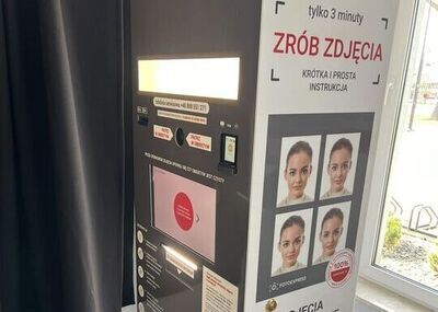 Kabina do robienia zdjęć dokumentowych FOTOEXPRESS z panelem sterowania i dużym ekranem oraz zasłoną po lewej stronie. Na górze jest jasny, podświetlany prostokątny panel. Pod nim widać instrukcje i elementy sterujące, w tym okrągły obiektyw aparatu pośrodku oraz mały czytnik/kasownik po prawej. Niżej znajduje się duży ekran dotykowy z różowym ekranem startowym i strzałką w prawo. Na prawym boku kabiny jest duża biała plansza z tekstem „tylko 3 minuty”, „ZRÓB ZDJĘCIA”, „KRÓTKA I PROSTA INSTRUKCJA” oraz przykładowymi czterema zdjęciami twarzy w układzie 2×2. Niżej jest nagłówek „ZDJĘCIA DOKUMENTOWE” i lista: „dowód osobisty, paszport, prawo jazdy, legitymacja, dyplom, wiza + inne”. Na dole widnieje napis „WERSJA CYFROWA GRATIS”. Kabina stoi przy oknie; na zewnątrz widać fragment chodnika i stojaki na rowery.