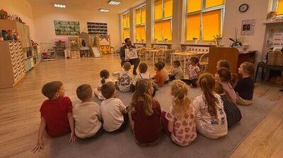 Sala przedszkolna z grupą małych dzieci siedzących w półkolu na dużym szarym dywanie. W środku, naprzeciw dzieci, klęczy dorosły mężczyzna w garniturze i trzyma otwartą książkę z obrazkami, jak podczas czytania lub prezentacji. Dzieci są zwrócone w jego stronę, większość siedzi na podłodze; część ma związane włosy w kucyki, ubrania są codzienne, w jasnych i kolorowych barwach.  Po prawej stronie znajdują się wysokie okna z intensywnie pomarańczowymi roletami, a pod oknami stoją rzędy małych stolików i krzeseł. Na ścianie po prawej wisi zegar. W głębi i po lewej stronie widać regały, półki i szafki z zabawkami oraz materiałami plastycznymi, a także kolorowe dekoracje i tablice z pracami lub ilustracjami. Podłoga jest drewniana, a na suficie są prostokątne lampy