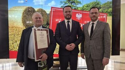 Trzech mężczyzn w garniturach stojących przed dużym ekranem z grafiką pola ze zbożem i belami siana oraz polskimi symbolami państwowymi.

W tle, w lewym górnym rogu, jest napis: „LUBELSKI URZĄD WOJEWÓDZKI w Lublinie” oraz biały orzeł.
Mężczyzna po lewej (starszy, jasna skóra, siwe włosy) trzyma w ramie dokument z dużym tytułem „DYPLOM”; na dokumencie widoczne jest imię i nazwisko zaczynające się od „Ryszard Li…”. Trzyma też ciemną torbę prezentową z zielonymi elementami i tekstem, którego nie da się w całości odczytać.
Mężczyzna pośrodku (jasna skóra, ciemne włosy, krótki zarost) ma granatowy garnitur i krawat; stoi z dłońmi złożonymi z przodu.
Mężczyzna po prawej (jasna skóra, krótkie ciemne włosy, zarost, okulary) ma szary garnitur i czarny krawat.

Na ekranie za nimi widać także dużą czerwoną planszę z białym orłem; część tekstu na niej jest zasłonięta i nieczytelna.