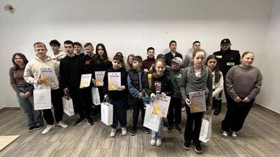 Grupa nastolatków i kilkorga młodszych dzieci ustawiona w dwóch rzędach przy białej ścianie w pustym pomieszczeniu z szarobrązową, drewnopodobną podłogą. Wiele osób trzyma białe torby prezentowe oraz dyplomy.

Po lewej stronie stoi dziewczyna w okularach, w brązowym swetrze i ciemnych spodniach. Obok niej chłopak w jasnej bluzie z kapturem trzyma dyplom; na dyplomie wyraźnie widać duży napis „DYPLOM”. Dalej w szeregu stoją głównie chłopcy w ciemnych bluzach i kurtkach, a kilka osób w tle jest częściowo zasłoniętych.

W środkowej części widać kilkoro młodszych dzieci z dyplomami „DYPLOM” i białymi torbami. Jedna dziewczynka z przodu, w ciemnej bluzie, też trzyma dyplom. Obok niej stoi chłopiec w zielonej bluzie z nadrukiem i czapce z daszkiem.

Po prawej stronie z przodu stoi dziewczyna w szarym szalu/kominie i ciemnych spodniach, trzyma dyplom „DYPLOM” oraz białą torbę. Najbardziej z prawej stoi dziewczyna w szaro-brązowej bluzie i ciemnych spodniach, z rękami splecionymi z przodu.

W tle po prawej stronie stoi dorosły mężczyzna w czarnym mundurze z naszywką „STRAŻ” i czarną czapką. Na ścianie wysoko po lewej widać małą, nieregularną plamę lub ubytek.