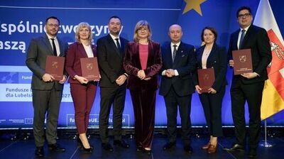 Grupa siedmiu osób stojących na scenie na tle niebieskiej ścianki z motywami Unii Europejskiej. To zdjęcie z wydarzenia oficjalnego.

Od lewej do prawej stoją cztery osoby trzymające bordowe teczki/dyplomy z herbem: mężczyzna w szarym garniturze i okularach, kobieta w bordowym garniturze, kobieta w granatowym żakiecie oraz mężczyzna w ciemnym garniturze i okularach (ten po prawej). Trzy osoby w środku nie trzymają teczek: mężczyzna w ciemnym garniturze, kobieta w bordowym welurowym komplecie z różową bluzką, oraz łysy mężczyzna w ciemnym garniturze.

Na tle widać dużą żółtą gwiazdę (symbol UE) i fragmenty napisów po polsku, m.in. „Fundusze … dla Lubels…” oraz „Dofinansowane przez Unię Europejską”. Po prawej stronie stoi flaga w barwach białej i czerwonej z herbem. Oświetlenie sceniczne jest mocne, z widocznymi lampami nad osobami.