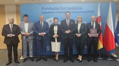 Grupa siedmiu osób w strojach formalnych stojąca w jednym rzędzie na tle niebieskiej ścianki informacyjnej. Na górze ścianki widoczny jest duży napis: „Fundusze Europejskie”, a poniżej mniejszy: „wspólnie tworzymy naszą przyszłość”. Po prawej stronie kadru stoją trzy flagi na masztach: flaga województwa warmińsko-mazurskiego (czerwono-żółto-biała z herbem), flaga Polski (biało-czerwona) oraz flaga Unii Europejskiej (niebieska z żółtymi gwiazdami).

Osoby ustawione są frontalnie, większość ma dłonie złożone przed sobą. Wszyscy wyglądają na dorosłych; sześciu mężczyzn i jedna kobieta. Mężczyźni mają na sobie garnitury w odcieniach czerni, granatu i szarości, białe koszule oraz krawaty; jeden z mężczyzn jest bez krawata. Kobieta stoi mniej więcej w środku; ma jasną, kremową sukienkę do kolan z ciemnym żakietem i czarne buty. Mężczyzna po prawej stronie trzyma bordową teczkę/okładkę z herbem i białym napisem; część napisu jest czytelna jako „MARSZAŁEK WOJEWÓDZTWA WARMIŃSKO‑MAZURSKIEGO”.

Pomieszczenie wygląda jak sala konferencyjna: sufit z okrągłymi lampami, po lewej stronie ściana z jasnymi panelami/żaluzjami, na podłodze wykładzina.