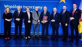 Grupa dziewięciu osób w formalnych strojach stoi na scenie przed niebieskim tłem z napisem „Wspólnie tworzymy…”. Jedna z osób trzyma dokument w czerwonej okładce. W tle widoczne elementy graficzne przypominające flagę Unii Europejskiej.