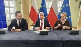 Trzy osoby przy stole podczas oficjalnego spotkania; w tle flagi Polski i UE, mężczyzna pośrodku podpisuje dokument.